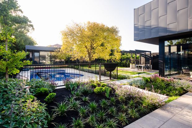 A landscaped modern residential courtyard with a swimming pool, black metal fencing, layered planting beds, paved walkways, and a contemporary home with dark glazing and geometric metal cladding. Outdoor dining and lounge areas are visible beside the building.