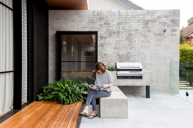 Contemporary courtyard outdoor living space with a timber-lined covered area opening onto a pale paved terrace. A concrete block feature wall incorporates a built-in stainless-steel barbecue, while a concrete bench, large leafy planting, and dark-framed glazing define the space.