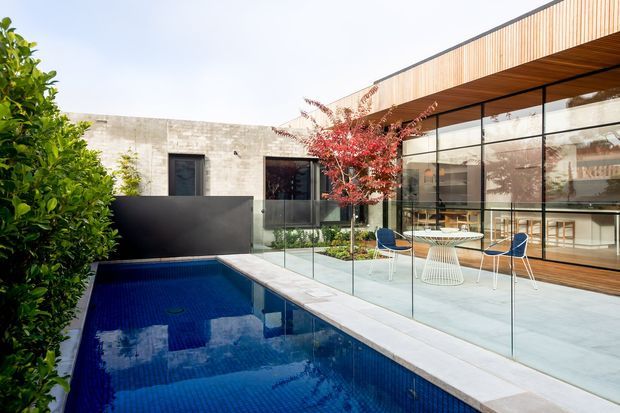 Contemporary courtyard-style residence with a narrow rectangular swimming pool, frameless glass balustrade, pale tiled terrace, and a minimalist outdoor dining area. The building combines a raw concrete masonry wall with extensive black-framed glazing and warm vertical timber cladding beneath a deep roof overhang. Lush planting and a red-leaf ornamental tree soften the modern architecture.
