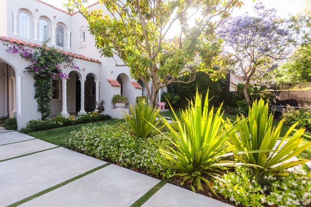 A landscaped courtyard garden in front of a white stucco residence with arched openings, shuttered windows, terracotta-tiled awnings, and climbing bougainvillea. Large pale concrete stepping slabs with narrow grass joints lead through manicured planting beds of spiky architectural plants, flowering groundcover, and mature trees.