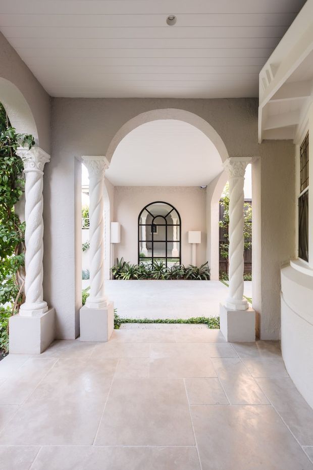 A refined covered entry porch or passage framed by white twisted classical columns and rounded arches. The space has pale stone-look tiled flooring, textured warm-grey stucco walls, a white tongue-and-groove ceiling, arched black-framed glazing, built-in shelving, and lush planting visible around the courtyard beyond.