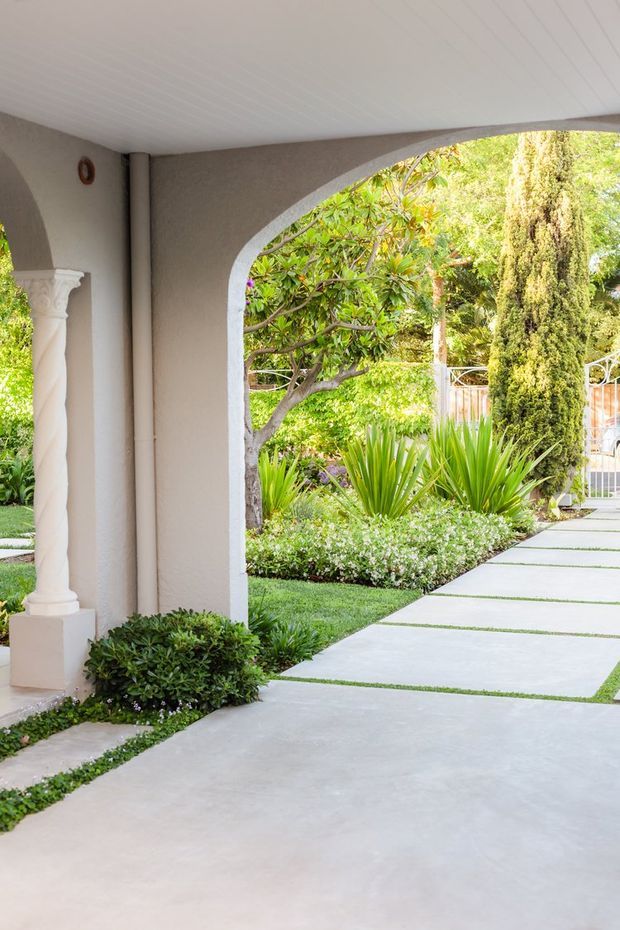 A covered exterior entry porch with a large rounded arch opening, pale stucco walls, a white paneled ceiling, and a decorative classical column. The view looks onto a landscaped garden with lush planting, clipped shrubs, and a light concrete stepping-paver walkway.