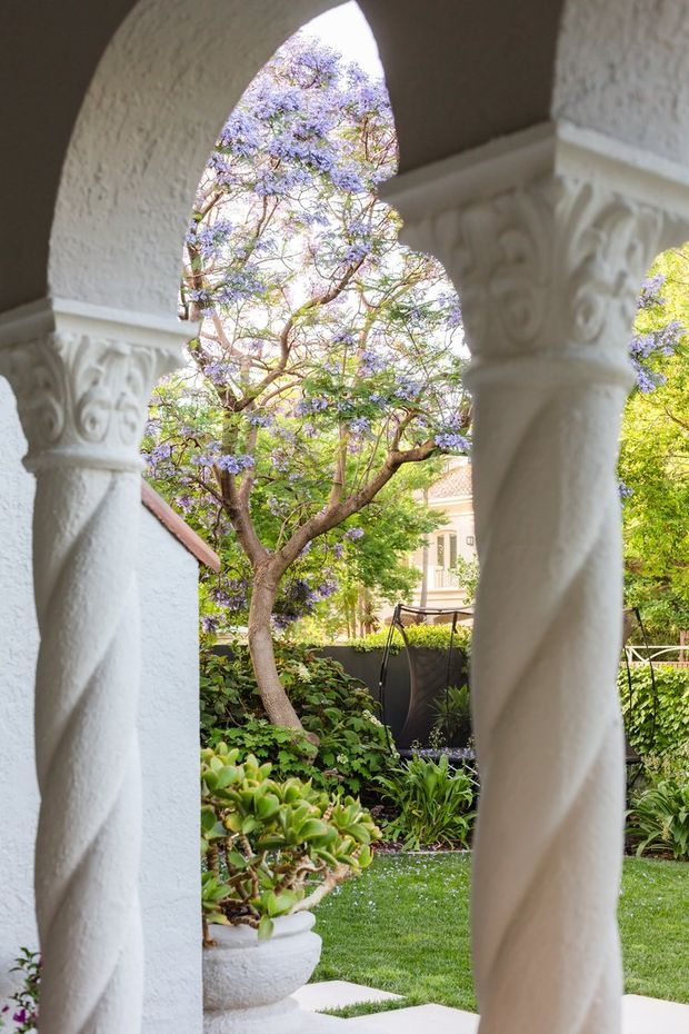 A view through a white stuccoed arched colonnade with ornate carved capitals and twisted columns, looking onto a lush courtyard garden with a flowering jacaranda tree, clipped lawn, planted borders, and a glimpse of a pale residential building beyond.