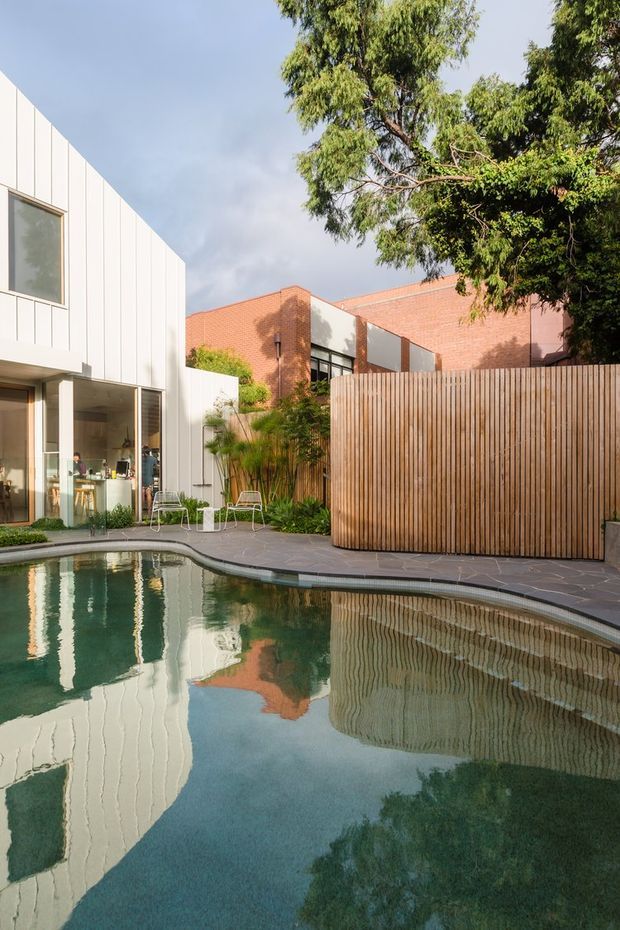 A landscaped residential courtyard with a curved swimming pool in the foreground. The pool reflects a contemporary white-panelled house and adjacent red-brick building forms. A tall timber privacy fence, paved pool surround, outdoor chairs, planted garden beds and mature trees complete the tranquil outdoor setting.