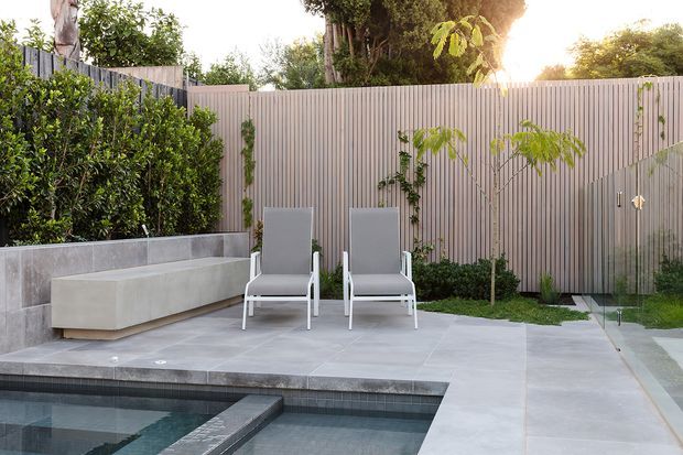 A contemporary landscaped courtyard with a compact rectangular swimming pool, pale grey tiled paving, two reclining chairs, and a built-in concrete bench. Vertical light-toned fencing provides privacy, while clipped hedges and young trees soften the hardscape. Warm sunlight creates a calm, refined outdoor setting.