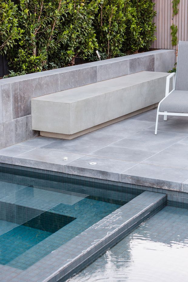 A contemporary outdoor pool area with geometric tiled water features, a large paved terrace, and a built-in concrete bench against a tiled retaining wall. Dense hedging provides greenery behind the seating area, with a partial outdoor lounge chair visible at right.