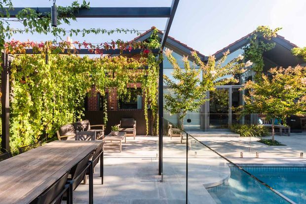 A sunlit landscaped courtyard and outdoor entertaining area with a long timber dining table, climbing greenery on brick and timber structures, a glazed pavilion, and a swimming pool in the foreground. The architecture combines pitched gable roofs, dark-framed glazing, timber elements, and exposed brick, creating a warm contemporary garden setting.