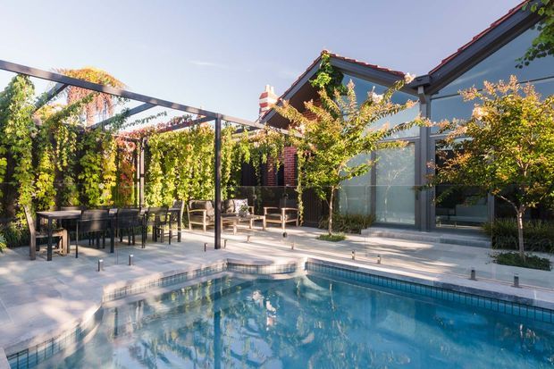 A sunlit courtyard pool area beside a contemporary glazed building. The pool is surrounded by pale paving, a dark metal pergola with lush climbing greenery, outdoor dining furniture, and small landscaped trees. The building combines large glass walls with dark-framed openings and a tiled pitched roof.