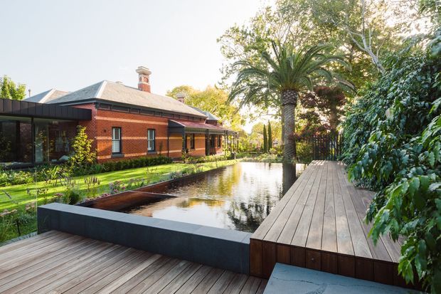 A landscaped courtyard beside a red-brick, single-storey heritage-style house, featuring a long reflecting pool or narrow swimming pool, timber decking, a raised timber boardwalk, mature planting, and a tall palm tree. The home has a low hipped metal roof, brick chimneys, dark-framed glazing, and a contemporary glazed extension at the left.