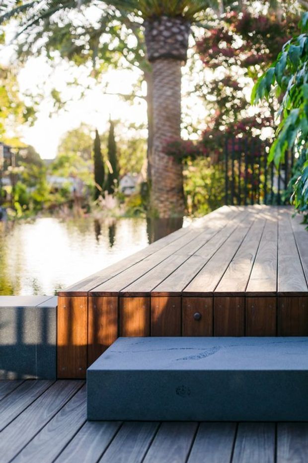 A sunlit timber deck beside a calm waterway, featuring a raised platform with warm brown timber-clad edging, built-in dark grey bench seating, and black metal railing amid lush tropical vegetation.