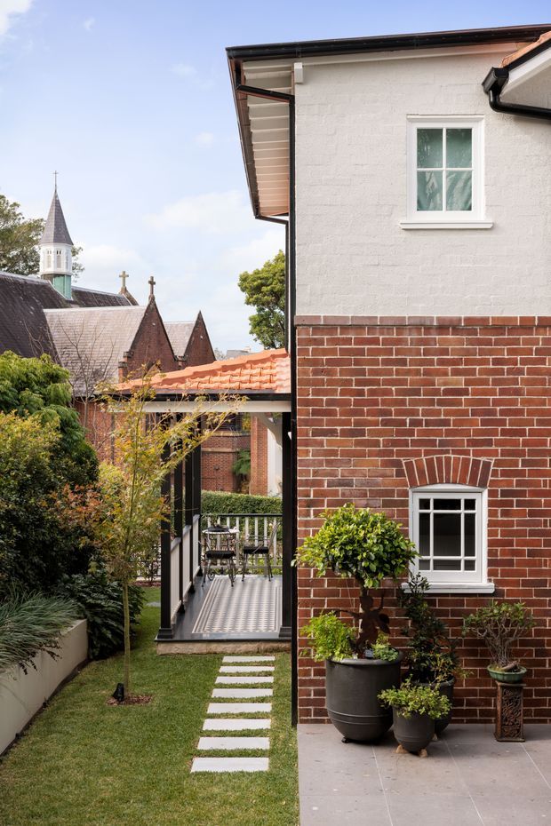 A landscaped courtyard passage beside a two-storey residential building, with a red brick lower facade, pale rendered upper wall, white-trimmed windows, and dark gutters. A narrow lawn with rectangular stepping stones leads to a covered outdoor deck with patterned tiled flooring and railings. Mature planting and potted topiary soften the space, while a historic church with pointed roofs and a small tower is visible beyond.