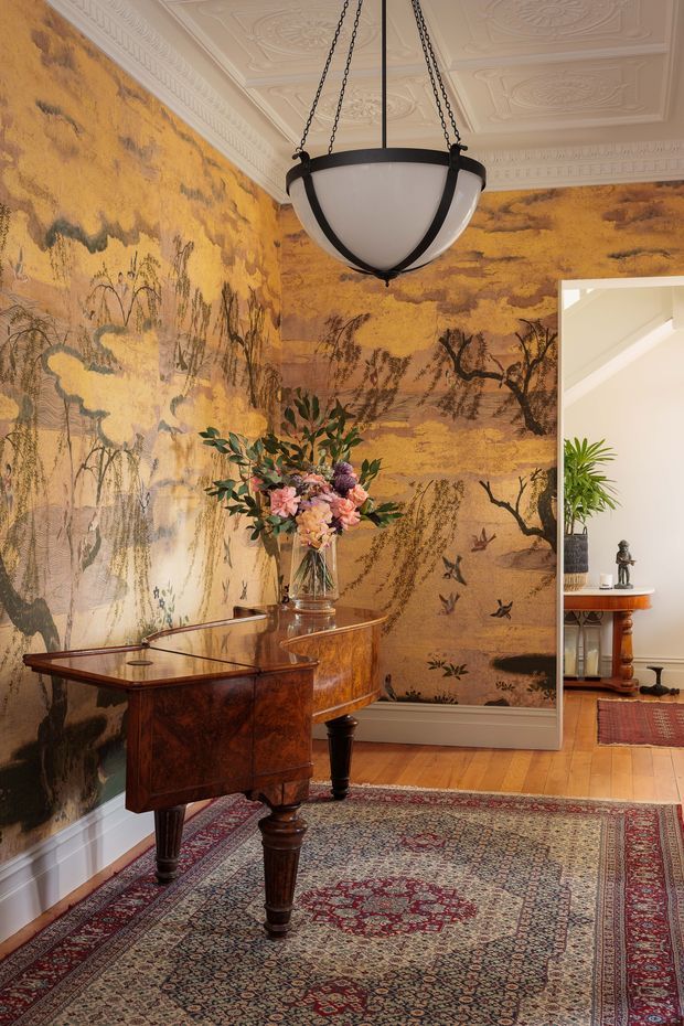 A richly decorated traditional interior corner featuring a dark polished timber console or writing desk, ornate patterned rug, dramatic ochre and brown scenic mural wallpaper, elaborate plaster ceiling medallions and cornices, and a large black-framed pendant lantern. A floral arrangement sits on the furniture, while an adjoining room is visible through the opening at right.