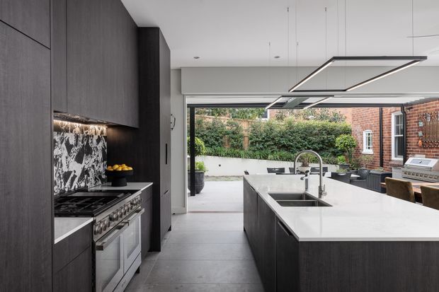 Contemporary kitchen with a large island, dark timber cabinetry, a pale stone benchtop, integrated cooking appliances, and a dramatic black-and-white stone splashback. Full-height glazing opens to a landscaped courtyard, while linear pendant lights and large-format grey floor tiles complete the refined monochromatic scheme.