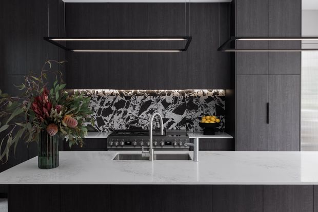 A sophisticated contemporary kitchen featuring dark vertical-grain cabinetry, a large pale stone island benchtop with an integrated sink and streamlined chrome tapware, and a dramatic black-and-white veined stone splashback. Linear pendant lights illuminate the island, while stainless-steel appliances and understated styling create a refined, high-contrast interior.