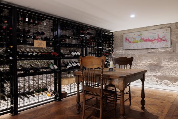A warmly lit residential wine cellar and tasting room with extensive black metal wine racks filled with bottles along the left wall, a rustic timber dining table and wooden chairs in the foreground, and textured pale stone walls. A framed wine-region map hangs above the table, while parquet timber flooring adds warmth and character.