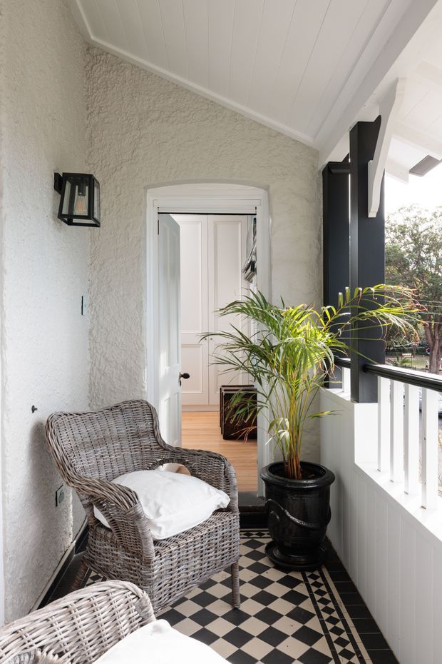 A bright covered veranda or entry porch with a black-and-white checkerboard tiled floor, textured light stucco walls, white beadboard-style ceiling and railing, and bold black structural posts. Wicker armchairs with white cushions sit beside a large potted palm. An open doorway leads into a light-filled interior with timber flooring and white paneled doors. A black wall lantern provides a decorative architectural detail.