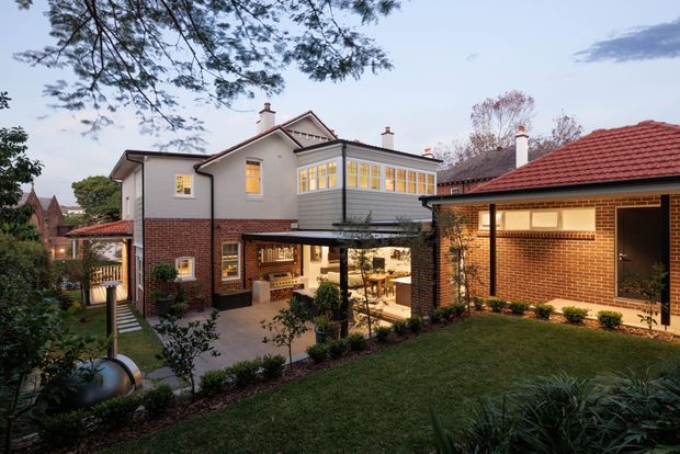 Exterior view of a multi-wing residential house arranged around a landscaped courtyard. The building combines red brick lower walls, light rendered upper walls, dark-framed glazing, pitched tiled roofs, gabled details and a glazed projecting room. A covered outdoor dining or entertaining area opens onto the courtyard, with established planting and lawn in the foreground.