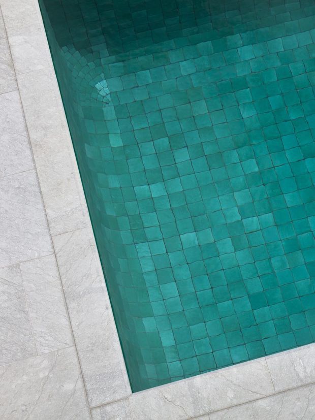 Close-up, top-down view of a swimming pool corner lined with small turquoise-green square tiles, bordered by large pale grey stone paving slabs. The image emphasizes the pool finish and clean geometric edge detailing.