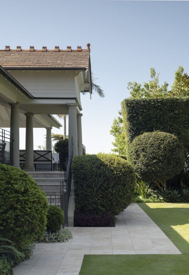 A landscaped residential exterior with a covered front veranda supported by classical columns, a tiled pitched roof, and broad steps leading to a pale tiled garden path. Carefully clipped hedges, rounded shrubs, lawn, and mature planting frame the entrance.