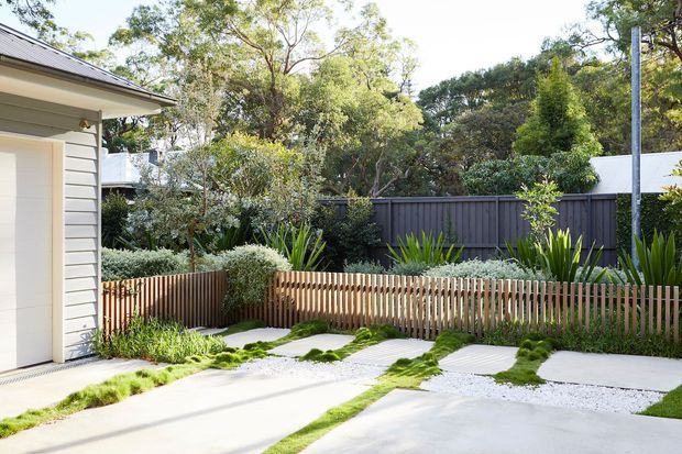 A bright landscaped residential driveway beside a light-coloured weatherboard building. The foreground features broad concrete parking or access strips separated by white gravel and narrow planted grass bands, with a low vertical timber picket fence, dense greenery, and a dark boundary fence behind.