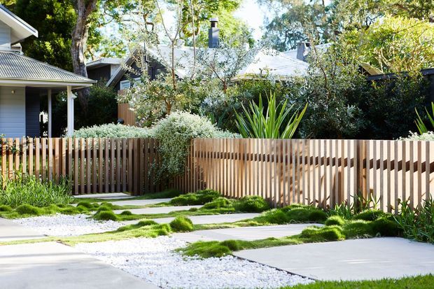 A landscaped residential front garden and walkway with pale concrete stepping slabs set through white gravel and low mounding groundcover. A vertical timber slat fence encloses a leafy garden, with single-storey houses featuring dark metal pitched roofs visible behind it. The scene has a contemporary, natural suburban character with strong greenery and timber detailing.