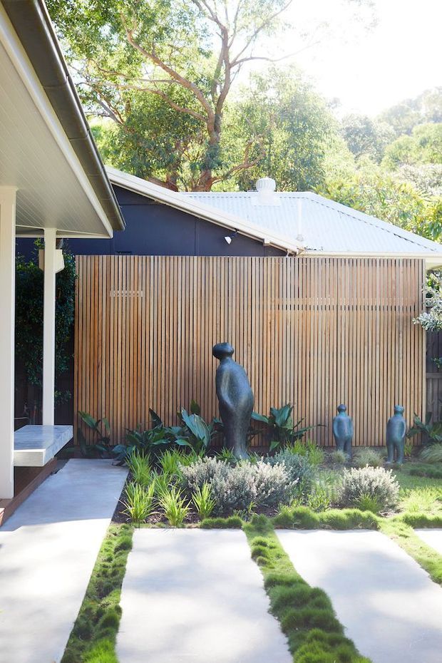 A landscaped courtyard or front garden beside a single-storey dwelling, featuring large pale concrete stepping slabs separated by strips of grass, low ornamental planting, sculptural figures, and a vertical timber batten privacy screen. The adjacent building has dark painted walls, a light metal pitched roof, and a covered entry with white posts and soffit.