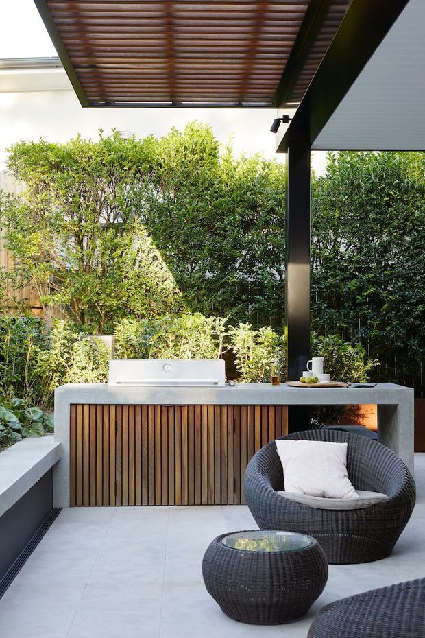 A refined outdoor entertaining area with a built-in barbecue kitchen beneath a pergola. The kitchen features a concrete counter and vertically slatted timber cabinetry, surrounded by lush screening plants. Woven dark outdoor lounge chairs and a low table sit on a pale tiled terrace, with strong black structural posts and timber pergola battens overhead.