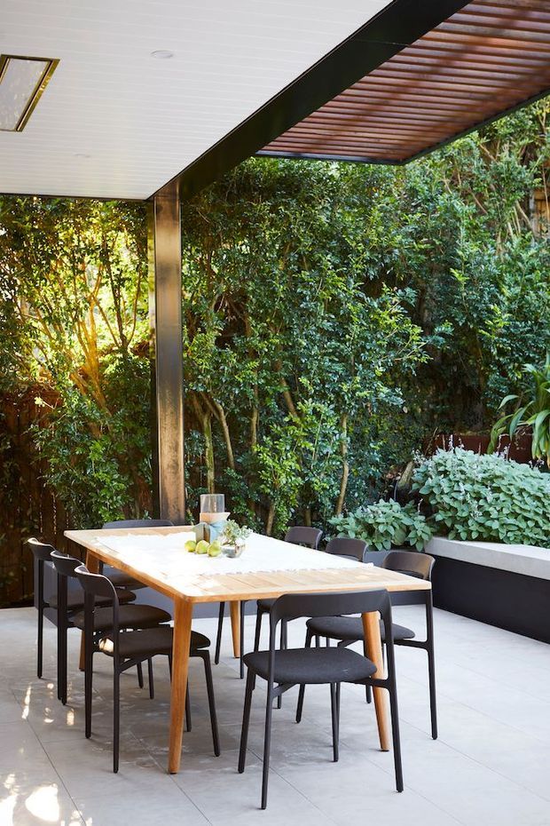 A covered outdoor dining area set within dense lush planting. A large light timber dining table with black chairs sits on pale tiled flooring, beneath a white paneled ceiling and a slatted timber pergola roof supported by dark structural posts. Raised black-edged planter beds frame the garden setting.