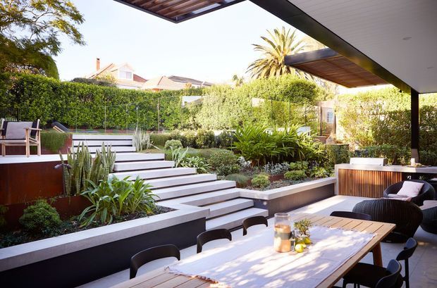 A refined outdoor entertaining area opening onto a landscaped courtyard garden. A large dining table sits beneath a deep roof overhang with timber-lined soffits, beside a lounge setting and built-in outdoor cabinetry. Broad stepped terraces with crisp light edging, dark retaining walls, layered planting, clipped hedges, and a water feature create a structured contemporary garden.