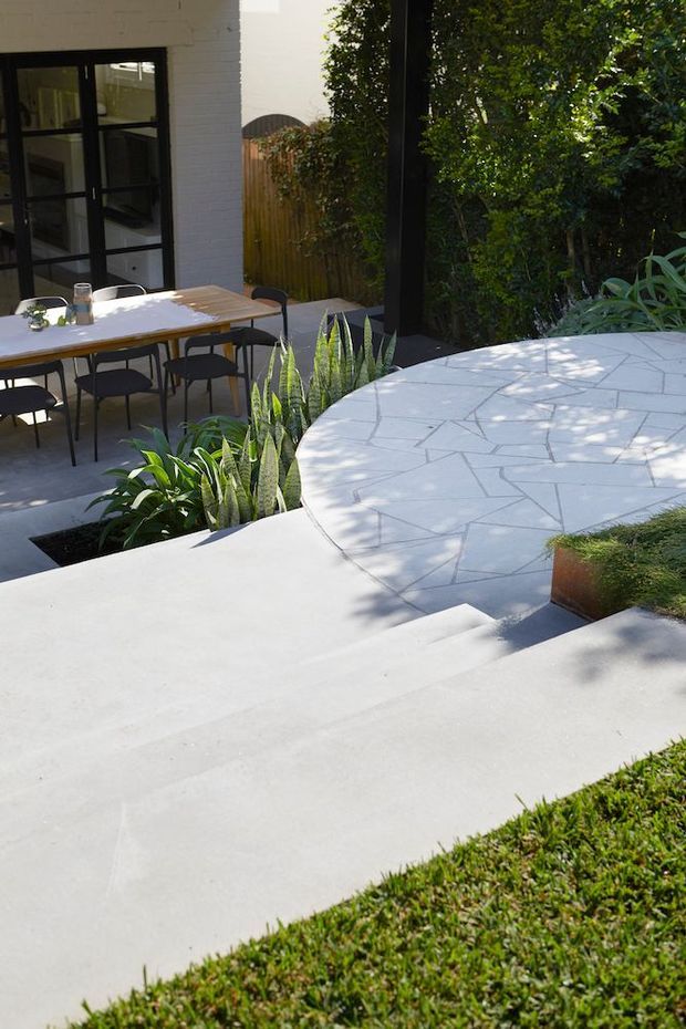 A sunny landscaped courtyard or garden terrace with broad pale concrete steps and a curved, irregularly patterned paved platform. A timber outdoor dining table with dark chairs sits beside a white brick building with black-framed glazed doors. Dense green planting, raised planters, and a timber privacy fence surround the outdoor space.