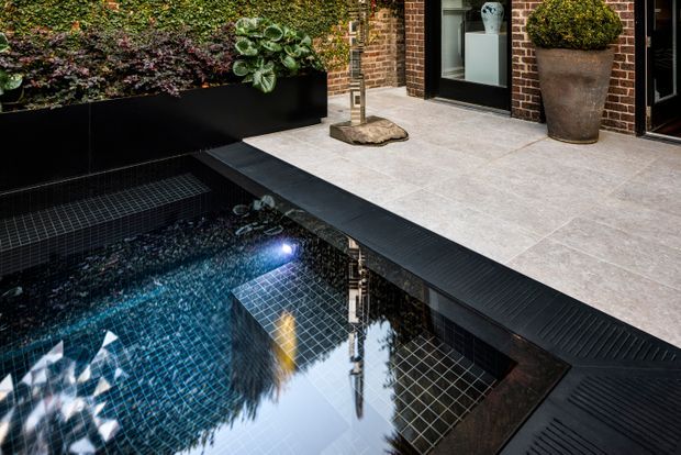 A compact courtyard pool beside a brick-clad building, featuring dark blue mosaic pool tiles, integrated underwater lighting, a tiled pool step or bench, and a pale stone-paved terrace edged with black drainage grilles. Raised black planters with dense greenery and large potted shrubs frame the space.