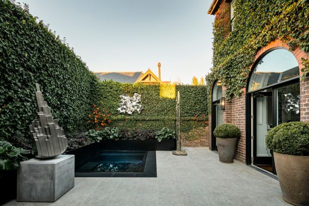 A secluded landscaped courtyard enclosed by dense clipped ivy-covered walls, with a small black-lined reflecting pool, sculptural planting, a contemporary metal sculpture on a concrete plinth, and a brick facade featuring a tall arched glazed doorway. Large clipped topiary pots and pale grey paving create a refined, private outdoor setting.