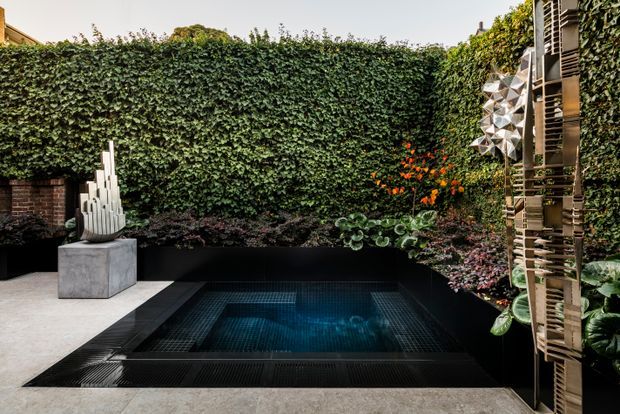 A compact landscaped courtyard with a dark tiled plunge pool or spa set into pale paving. Dense ivy-covered boundary walls create a private green backdrop, with layered planting, orange flowers, a sculptural artwork on a concrete plinth, and a timber slatted screen along the right side.