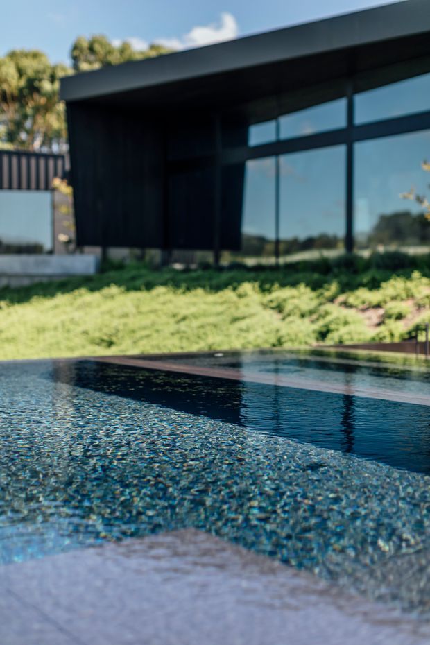 A modern dark-clad house with expansive black-framed glazing is viewed across a reflective swimming pool. The pool occupies the foreground, with a pale paved edge and landscaped lawn behind it. The building has a strong horizontal, low-slung form and a flat roof, while sunlight and reflections create a calm outdoor residential setting.