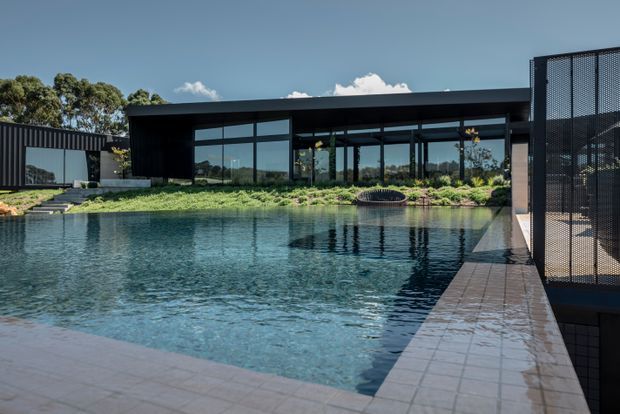 Contemporary single-storey pavilion with dark cladding, extensive floor-to-ceiling glazing and a flat roof, positioned beside a large reflective swimming pool. The landscaped grounds include low planting and a curved outdoor feature, while a tiled pool edge and perforated metal-screen structure frame the foreground.