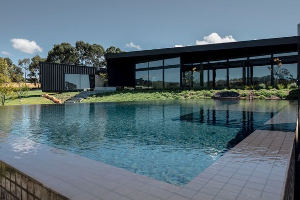 Wide exterior view of a sleek, low-profile modern residence with extensive dark cladding and floor-to-ceiling glazing, set behind a large swimming pool. The pool is surrounded by tiled paving and landscaped garden beds, with a separate dark-clad pavilion or wing to the left. The image has a crisp, contemporary architectural character and strong reflections in the water and glazing.
