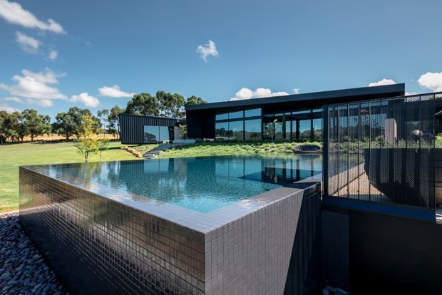 A modern single-storey residence set in an open landscaped site, featuring a large raised swimming pool with dark tiled sides and surrounding terrace. The building has a long low profile, extensive floor-to-ceiling glazing, dark metal cladding and a screened outdoor structure. Clear blue sky and lawn create a bright, contemporary architectural setting.
