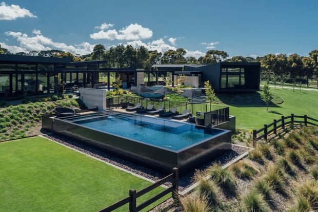 A wide aerial view of a contemporary single-storey property set within landscaped grounds. The dark low-profile building has extensive glazing and several connected wings arranged around a central outdoor entertaining area. A raised rectangular swimming pool with integrated lounge decking dominates the foreground, bordered by ornamental grasses, manicured lawn, fencing, and garden planting.