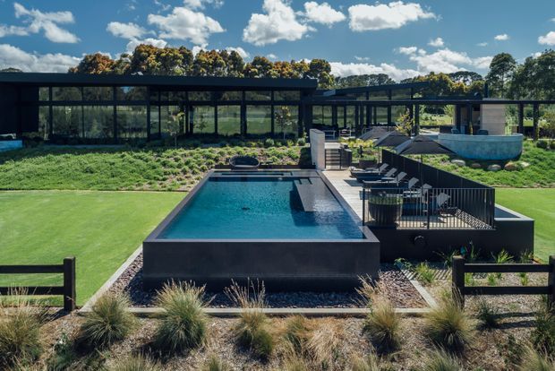 A landscaped outdoor pool area viewed from the garden, featuring a rectangular dark-edged swimming pool, raised deck with outdoor seating, lawn, ornamental grasses, and a long low modern pavilion with extensive black-framed glazing in the background. A curved raised spa or water feature is visible to the right under a bright blue sky.