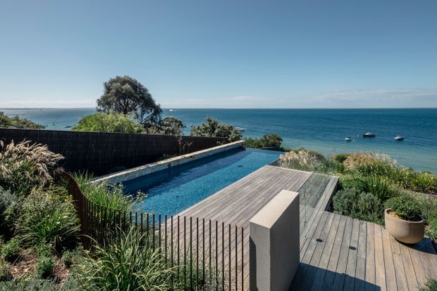 A contemporary coastal outdoor entertaining area overlooking the ocean, featuring a long narrow swimming pool bordered by timber decking, dark privacy fencing, native ornamental planting, and sculptural concrete screening. The view includes a calm blue sea and small boats beyond the landscaped property.