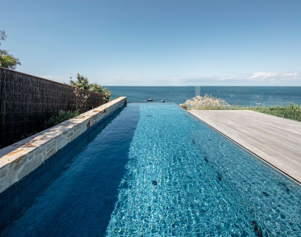 An infinity-edge swimming pool extends toward a broad ocean horizon. The pool is bordered by a pale stone wall and dark privacy screening on the left, with a timber deck and coastal planting on the right. The scene is bright, open, and strongly connected to the seafront.