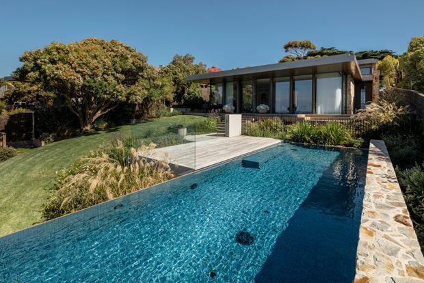 A modern single-storey residence set within a landscaped garden, viewed from an expansive blue swimming pool. The house features extensive floor-to-ceiling glazing, a broad low-pitched roof, dark structural framing, and a timber-toned terrace. A stone-clad pool edge and planted garden border the pool, creating a refined indoor-outdoor setting.