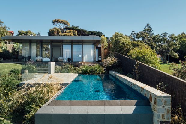 Contemporary single-storey residence set in a landscaped suburban setting, with a flat roof, extensive floor-to-ceiling glazing, dark exterior framing, and a narrow rectangular swimming pool extending toward the foreground. A raised stone-clad retaining wall and timber-toned boundary fence define the pool terrace, while layered planting surrounds the building and pool.