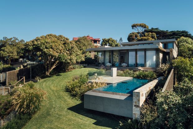 A landscaped residential property viewed from the garden, featuring a modern single-storey flat-roof pavilion with extensive floor-to-ceiling glazing, a lawn, planted garden beds, and a raised rectangular swimming pool with a stone-clad edge and tiled terrace. Mature trees and neighbouring residential structures are visible in the background.