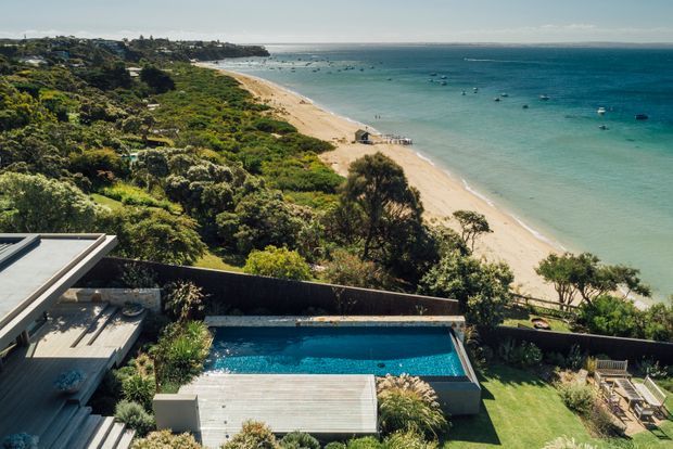 Elevated coastal property overlooking a sandy beach and turquoise bay, with a rectangular swimming pool, timber deck, landscaped garden, and portions of a contemporary flat-roofed residence in the foreground.