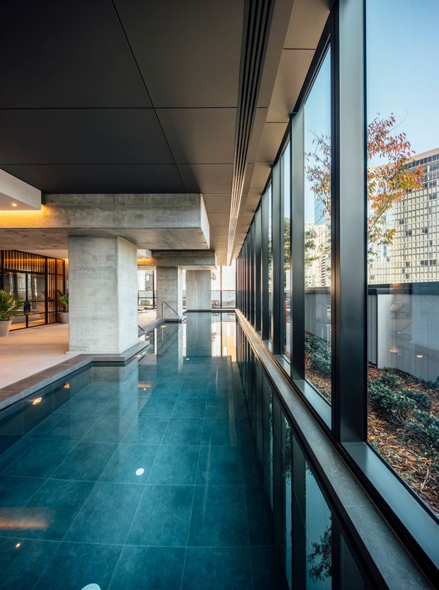 A sophisticated indoor pool corridor in a contemporary commercial or residential building. The long space features glossy dark teal floor tiles, a reflecting pool or water feature along the left side, exposed concrete columns and beams, a dark paneled ceiling, warm integrated lighting, and full-height black-framed glazing overlooking landscaped planting and adjacent urban buildings.
