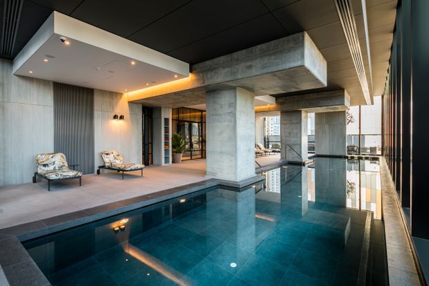 A dramatic modern indoor pool area with a deep turquoise tiled pool, exposed concrete columns and beams, dark ceiling panels, full-height glazing, and a raised lounge platform with two chairs. Warm recessed and concealed lighting contrasts with the cool concrete and water, creating a luxurious, architectural atmosphere.