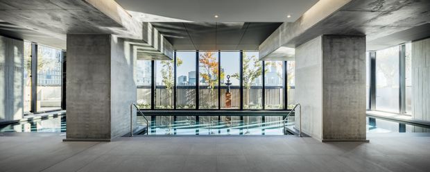 A sophisticated indoor pool area with a long rectangular swimming pool, polished concrete columns and ceiling beams, extensive floor-to-ceiling glazing, and dark metal balustrades. The space has a strong exposed-concrete architectural character, abundant natural light, and views toward an urban setting and trees outside.