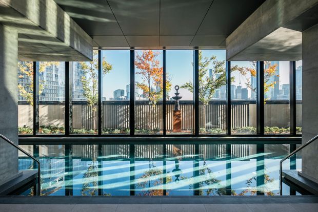 A dramatic indoor swimming pool viewed from the pool steps, framed by floor-to-ceiling black-framed glazing. Exposed concrete beams and columns create a robust modern architectural character, while the pool reflects the city skyline, trees, and autumn light outside. A decorative pedestal stands beyond the glazing on a planted terrace.