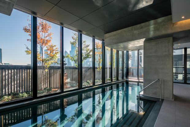 A contemporary indoor swimming pool within a concrete-framed building, featuring floor-to-ceiling glazing, dark metal window frames, a dark ceiling, and a tiled pool surround. Reflections of trees and the urban skyline are visible through the windows and across the water.